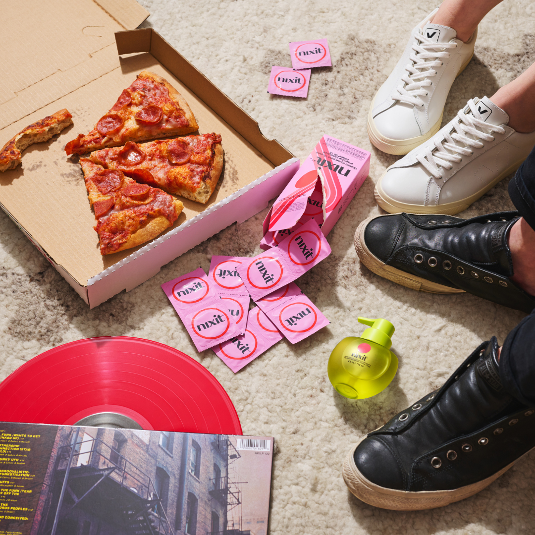 Open pizza box with pepperoni pizza, scattered pink packets, two pairs of sneakers, red vinyl record, and green bottle on a beige carpet.