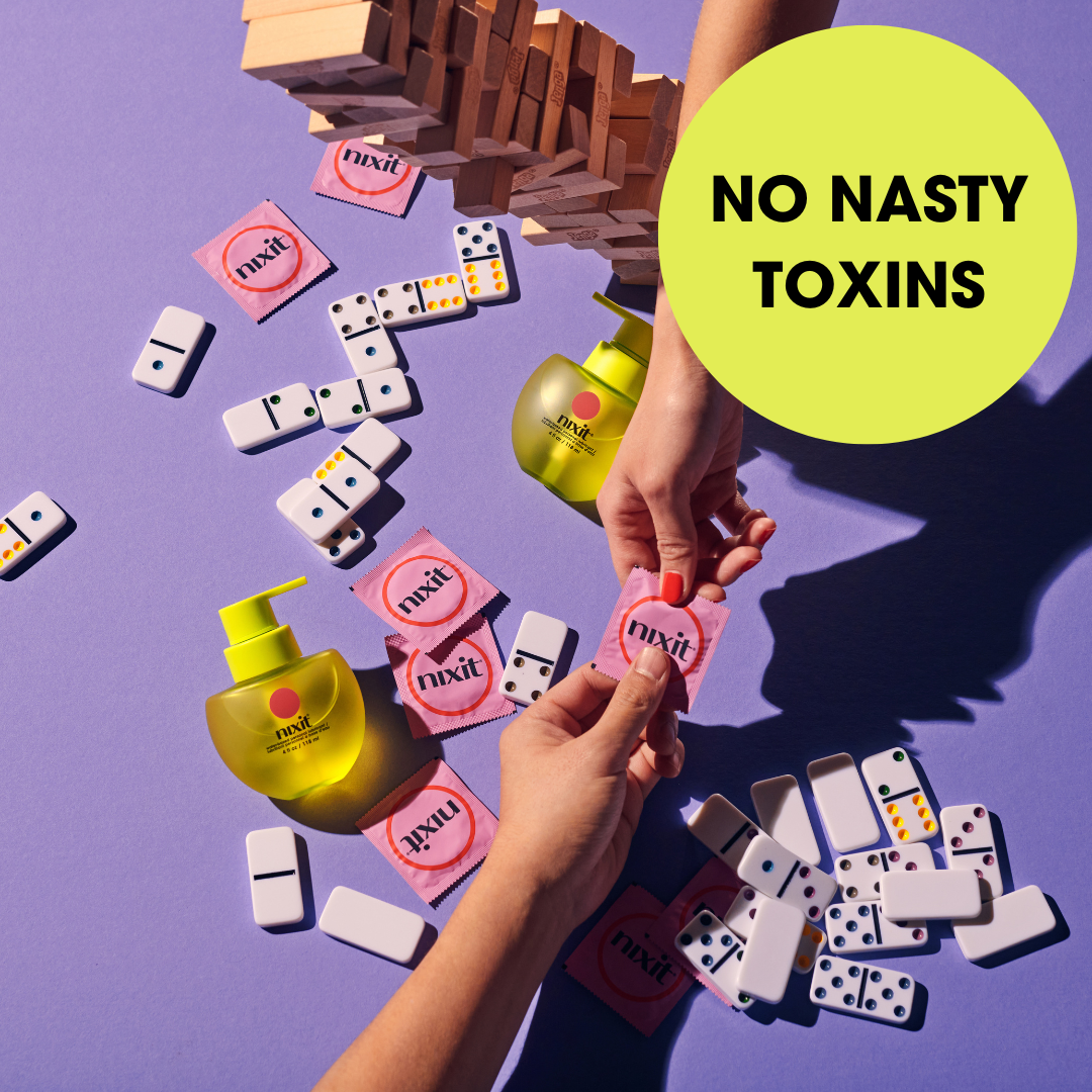 two arms extend over a messy table of dominos to exchange a condom in it's wrapper. On the table are nixit condoms and a wooden block game, with a bottle of nixit personal lubricant. Wording in a circle says 'no nasty toxins'