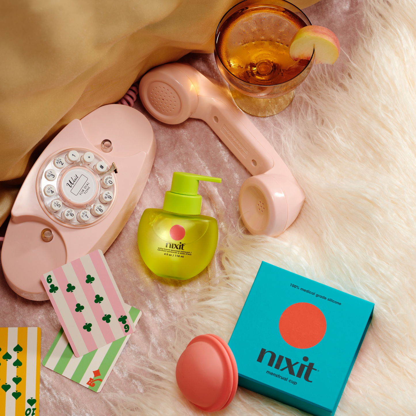 a nixit menstrual disc lies on a soft carpet next to the nixit water-based personal lubricant, with a phone, playing cards and a drink.